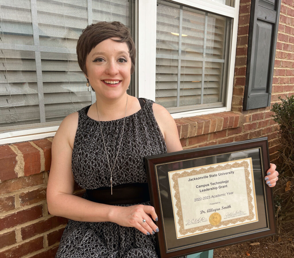 Allegra, a white woman with short brown hair and winged eyeliner, sits in front of a brick wall and poses with a framed certificate for her 2022-2023 Camus Technology Leadership Grant at Jacksonville State University