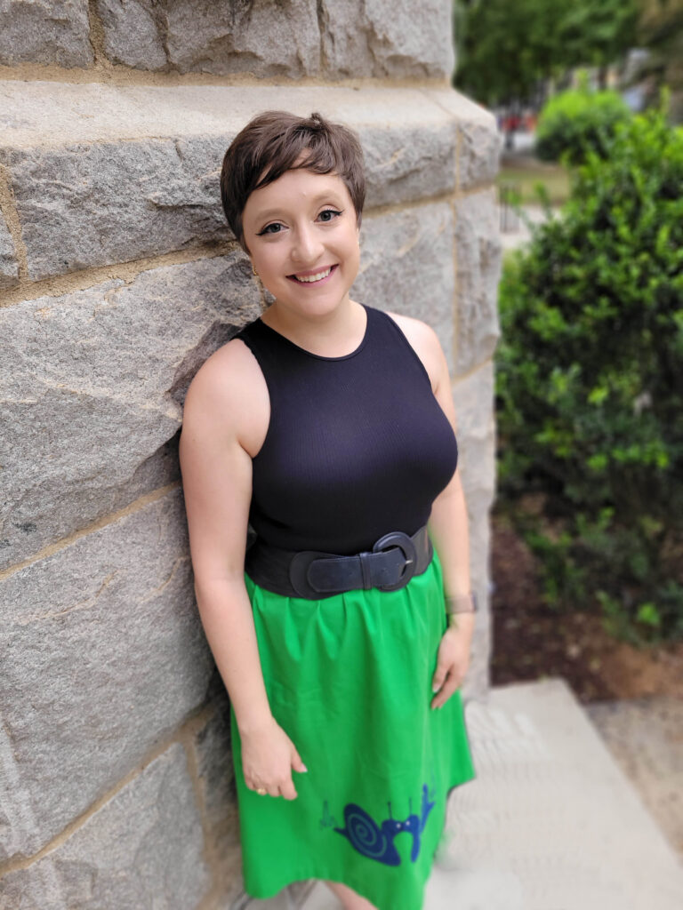 Allegra, a short white-woman with close-cropped brown hair, grins in front of a stone wall